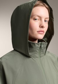 Gevoerde jas van gedempt groene stof, met een ritssluiting, een gladde textuur en schuine naaddetails voor een getailleerd silhouet.