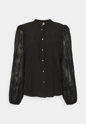 Blusa negra con frente abotonado, cuello redondeado y mangas abullonadas de bordado transparente. La textura varía entre sólida y encaje.