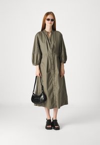 Olive roheline, põlvepikkune nööbid üles dress, millel on balloonvarrukad ja tikkimistooted. Must ristiõla kott ja jämedad mustad sandaalid.