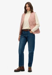 Un gilet matelassé rose sur un col roulé crème, associé à un jean bleu et des bottes à lacets marron. Il présente un devant à boutons et des poches.