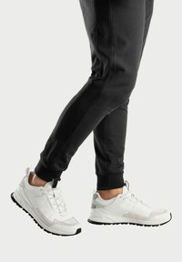 Weiße Sportsneaker aus weichen Materialien, mit Schnürdesign, grauen Akzenten und schwarzer Gummisohle. Kombiniert mit schwarzen Jogginghosen.
