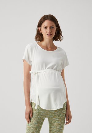 Witte zwangerschapsblouse met korte mouwen, een ronde halslijn en een zijstrikaccent; gedragen met gestreepte groen en gele textuur broek.