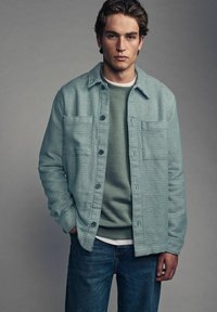 Light blue patterned shirt with two chest pockets, layered over a green sweatshirt, worn with blue jeans. Textured fabric and button details.