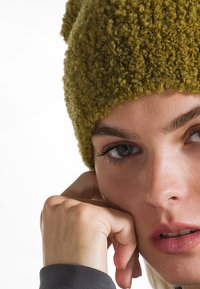 Primer plano de una persona con un gorro de punto verde texturizado, descansando su barbilla sobre una mano, con rasgos faciales visibles y tono de piel clara.