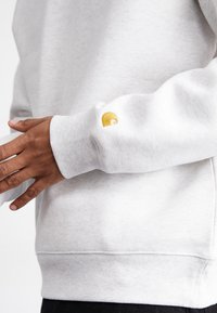 Lysegrå sweatshirt med ribbede manchetter og kant. Har et lille gult broderet logo på venstre ærme. Blødt, struktureret stof.