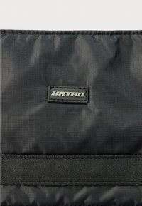 Μαύρη τσάντα από νάιλον με υφή, ορθογώνιο ταμπελάκι με λογότυπο σε μαύρο και ασημί και οριζόντια υφασμάτινη τιράντα.