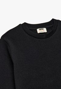 Schwarzer Waffelstrickpullover mit rundem Ausschnitt und weicher Textur. Zeigt ein Etikett mit der Aufschrift "Koton Kids" und Größenangaben.