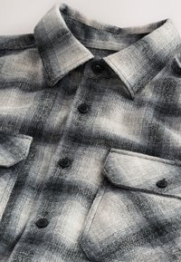 Camicia di flanella in tonalità grigie con motivo a quadretti neri e bianchi, caratterizzata da scollo con colletto, chiusure con bottoni e due tasche sul petto.