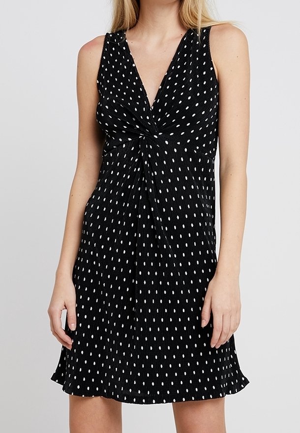 Robe noire sans manches à pois ovales blancs, col en V et détail de nœud torsadé à l'avant, portée par une femme debout.