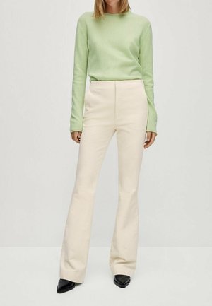 Kvinde iført lys grøn langærmet sweater, højtaljede cremefarvede bukser med vide ben og sorte spidse sko, stående mod en ensfarvet baggrund.
