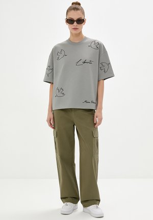 Donna che indossa una t-shirt grigia oversize con illustrazioni di uccelli e testo "Liberté", pantaloni cargo verdi oliva, sneakers bianche e occhiali da sole scuri.