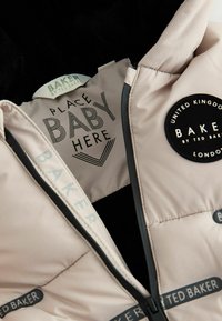 Ge polsterter Babytrage in hellrosa Stoff mit schwarzem Fleece-Futter, mit dem Text "BABY HIER PLATZIEREN" und einem runden Logo "BAKER BY TED BAKER".