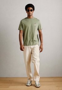 Olijfgroene t-shirt met korte mouwen en een kleine grafische print, gecombineerd met crèmekleurige broek en grijs-groene sneakers. Soepele stof.