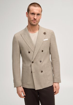 Man draagt een beige dubbelgeknoopt colbert met een bloemspeld op de revers, licht overhemd en donkere broek tegen een effen achtergrond.