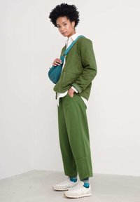 Grön vadderad jacka över en vit skjorta, tillsammans med matchande gröna byxor. Accessoarer inkluderar en blå axelväska och vita sneakers.