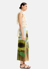 Beżowy top bez rękawów z drapowanym detalem; plisowana spódnica w odcieniach zieleni i brązu z nadrukiem słoneczników; czarne sandały na ramiączkach.