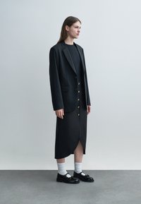 Γυναίκα που φοράει μαύρο σακάκι, μαύρη φούστα με κουμπιά, λευκές κάλτσες και μαύρα παπούτσια, στέκεται μπροστά σε έναν απλό ανοιχτό γκρι φόντο.