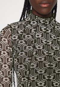 Tissu noir transparent comportant un motif floral blanc répétitif. Encolure haute avec des plis et des accents de couture verts contrastants.