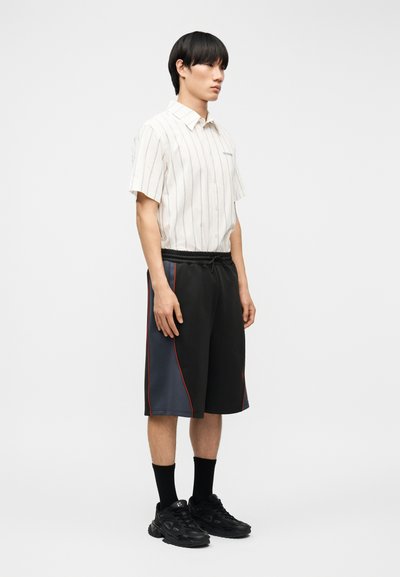 Άνδρας που στέκεται φορώντας ένα λευκό πουκάμισο με κοντά μανίκια και λεπτές ρίγες, μαύρο και ναυτικό σορτς με κόκκινη γραμμή, μαύρες κάλτσες και μαύρα αθλητικά παπούτσια.