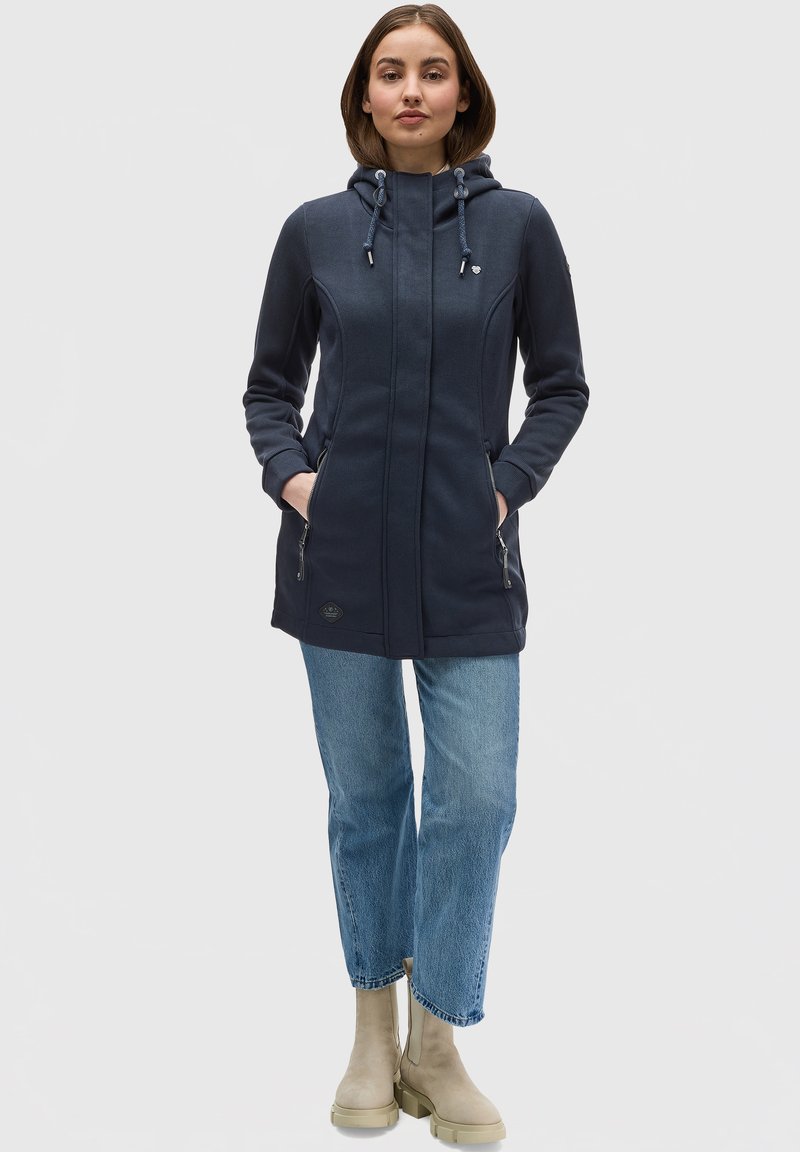 Námornícky modrá bunda s kapucňou a predným zipsom, dvomi postrannými vreckami a bielymi akcentmi. Kombinovaná s svetlomodrými džínami a béžovými členkovými čižmami.
