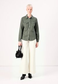 Camisa de manga longa verde-oliva com dois bolsos no peito, combinada com calças brancas de perna larga e uma bolsa preta estampada. Sapatos pretos de bico fino.