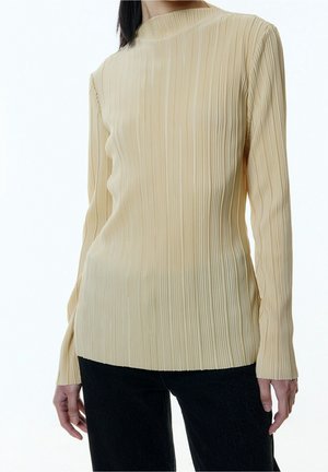 Femme portant un haut plissé beige à manches longues associé à un pantalon noir, debout devant un fond clair uni.