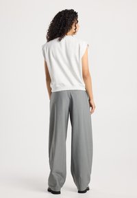 Woman with curly hair wearing a sleeveless light gray top and wide-leg gray trousers, standing and facing away on a white background.