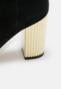 Chaussure à talon haut en daim noir avec un talon bloc doré côtelé et une base noire, détaillée par une texture lisse et des lignes épurées.