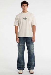 Beige katoenen T-shirt met een "ORIGINALS"-opdruk, gecombineerd met blauwe denim cargobroek met meerdere zakken en een verbleekte textuur.