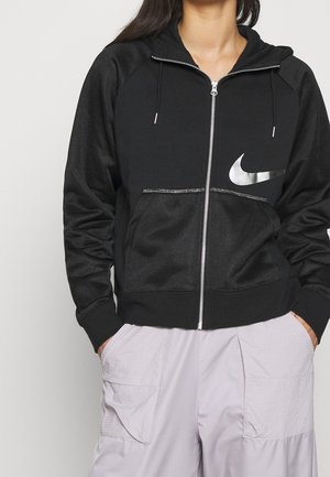 Person trägt schwarzen Nike-Kapuzenpullover mit Reißverschluss und silbernem Logo sowie hellgraue Cargohose, die Hände in den Taschen, gezeigt vom Hals bis zur Hüfte.