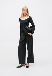 Μαύρο τοπ με μακριά μανίκια και φαρδιά λαιμόκοψη, συνδυασμένο με χαλαρά γκρι παντελόνια. Το μοντέλο κρατά μια μεγάλη μαύρη τσάντα pouch. Μαύρα παπούτσια με ψηλό τακούνι.