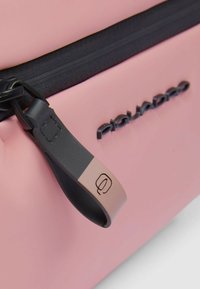 Bolsa de silicona rosa con cremallera negra y una lengüeta de cuero con un logo. Textura suave y diseño minimalista.