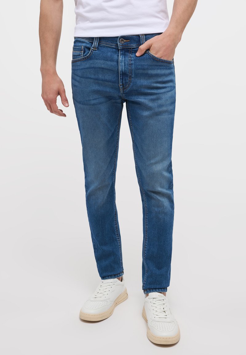 Vaqueros de denim azul con un corte ajustado, que cuentan con cinco bolsillos, un desvanecimiento sutil y un cierre clásico de botón. Combinados con zapatillas blancas.