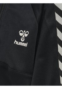 Svart sportjacka med vitt Hummel-logotyp och randigt mönster. Mjuk textur med dragkedjestängning och kontrasterande materialdetaljer.