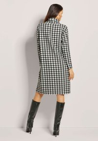 Schwarzes und weißes Hahnentristermuster, langärmliges Kleid mit hohem Kragen, kombiniert mit knielangen schwarzen Lederstiefeln.