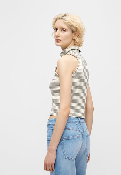 Νεαρή γυναίκα με κοντά σγουρά ξανθά μαλλιά, που φοράει αμάνικη ριγέ μπλούζα με λαιμόκοψη ζιβάγκο και ανοιχτόχρωμο τζιν, κοιτάζοντας πάνω από τον ώμο της.