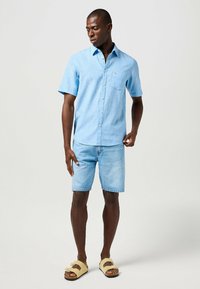 Camicetta azzurra a maniche corte con tasca, abbinata a pantaloni corti in denim chiaro e sandali beige, caratterizzata da una vestibilità comoda e un design informale.