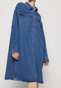 Denim shirt dress in blue, featuring a classic collar, two chest pockets, button front, long sleeves, and side slits at the hem.