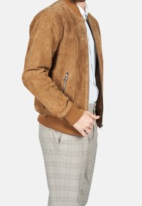 Man in een bruin suède bomberjack over een wit shirt en beige geruite broek, staand met een hand bij zijn zak tegen een witte achtergrond.