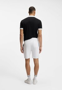 Hombre de pie de espaldas vistiendo una camisa polo negra con borde blanco, pantalones cortos blancos, calcetines blancos con texto negro y zapatillas blancas.