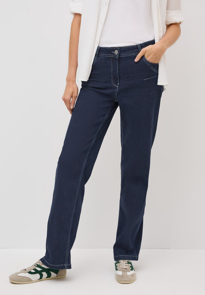 Jeans en denim bleu foncé avec une coupe droite, couture visible, poche avant, portés avec une chemise blanche et des baskets décontractées.