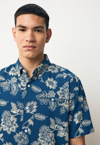 Camicia a maniche corte blu con bottoni, caratterizzata da un motivo floreale bianco, colletto strutturato e tessuto strutturato con chiusure a bottoni visibili.