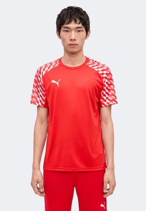 Tânăr care poartă un tricou sport roșu Puma cu modele geometrice albe pe mâneci și pantaloni scurți roșii asortați, stând în fața unui fundal simplu.
