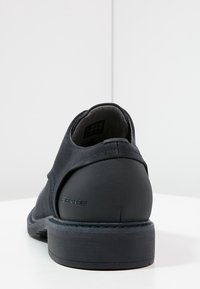 Chaussure bleu marine avec une texture lisse, talon arrondi et semelle en caoutchouc ; présente un dessus en tissu à motifs et un logo discret à l'arrière.