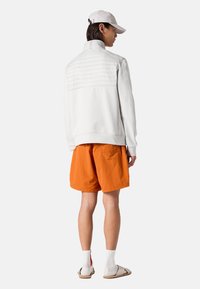 Weißer Sweatshirt mit strukturiertem oberen Abschnitt, kombiniert mit orangen Shorts, weißen Socken und Sandalen, von hinten gezeigt.