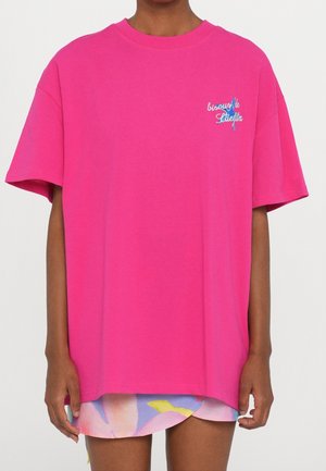 Osoba nosząca ogromną jasnoróżową koszulkę z małym niebiesko-białym napisem na lewej piersi, na kolorowej, kwiecistej spódnicy.