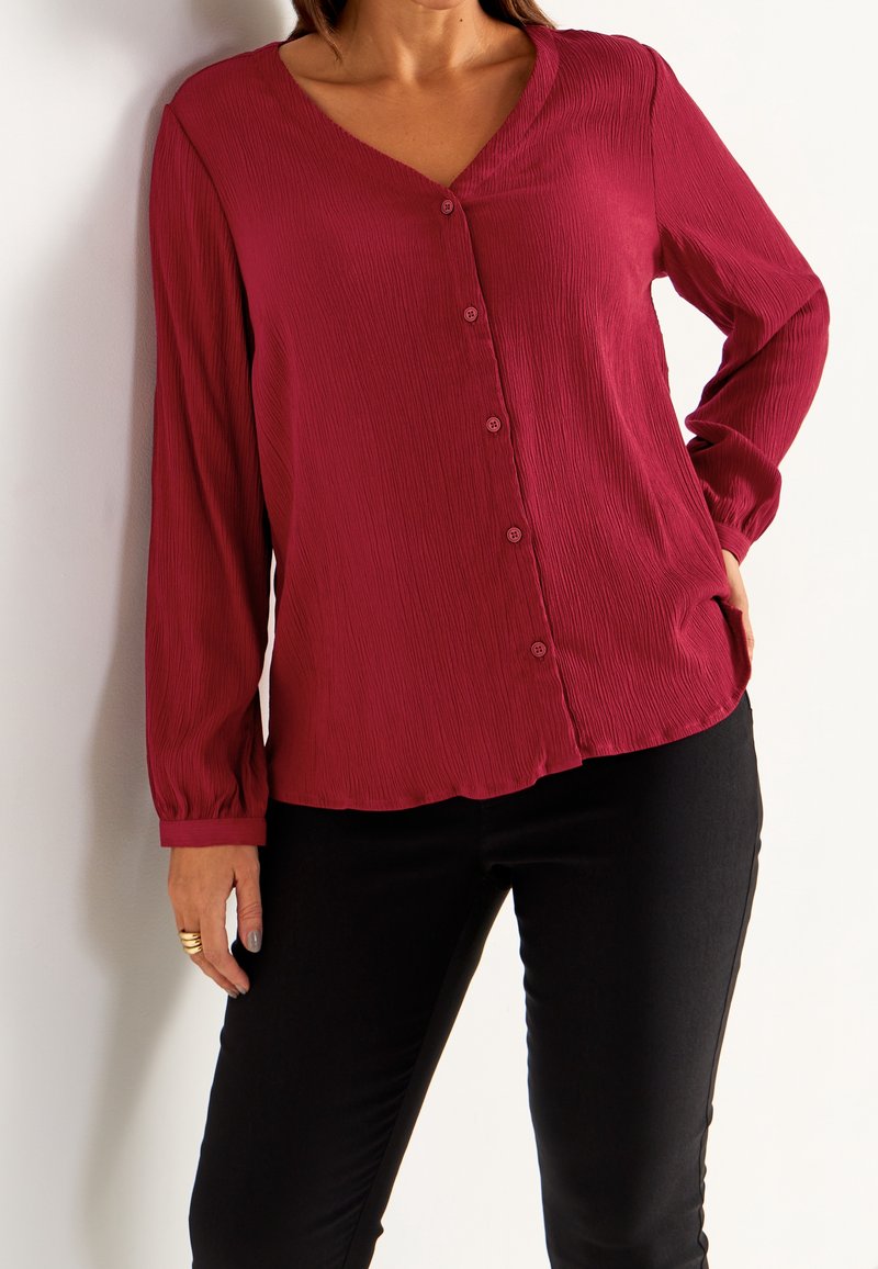 Blusa rossa con trama, scollatura a V, maniche lunghe e chiusura con bottoni. Presenta un orlo arrotondato e tasche laterali. Abbinata a pantaloni neri.