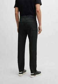 Pantalons noirs texturés avec une coupe ajustée, dotés de cinq poches et d'une ceinture de marque. Portés avec des baskets noires et une chemise noire.