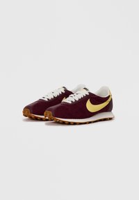 Raudoni Nike sportbačiai su geltonu swoosh ženkliuku, baltais raišteliais ir guminiu padu ant balto fono.