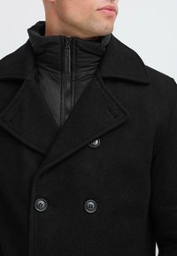 Schwarzer Woll-Peacoat mit doppelter Knopfleiste, großem Kragen und strukturierter Stoffqualität. Verfügt über glatte schwarze Reißverschlüsse und Knöpfe.